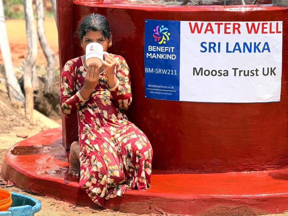 A young girl drinks clean water from a cup beside the finished red well and its Moosa Trust UK sign