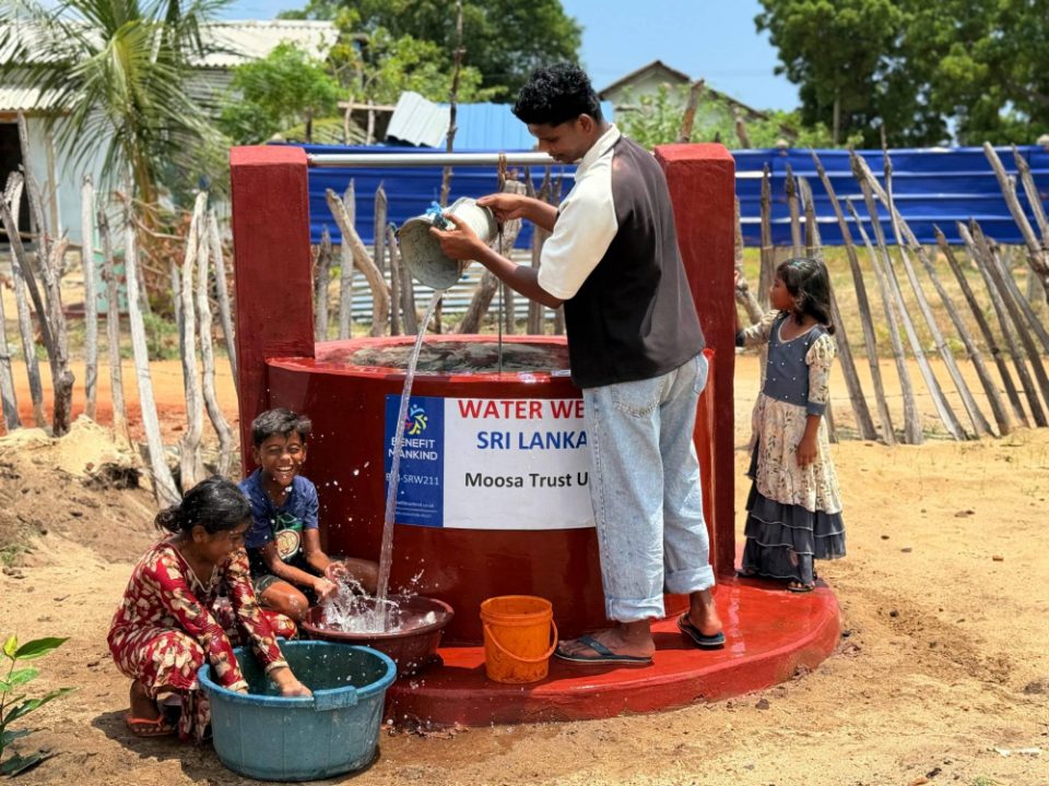 A young man pours a bucket of water from the new well while children laugh and splash in basins below